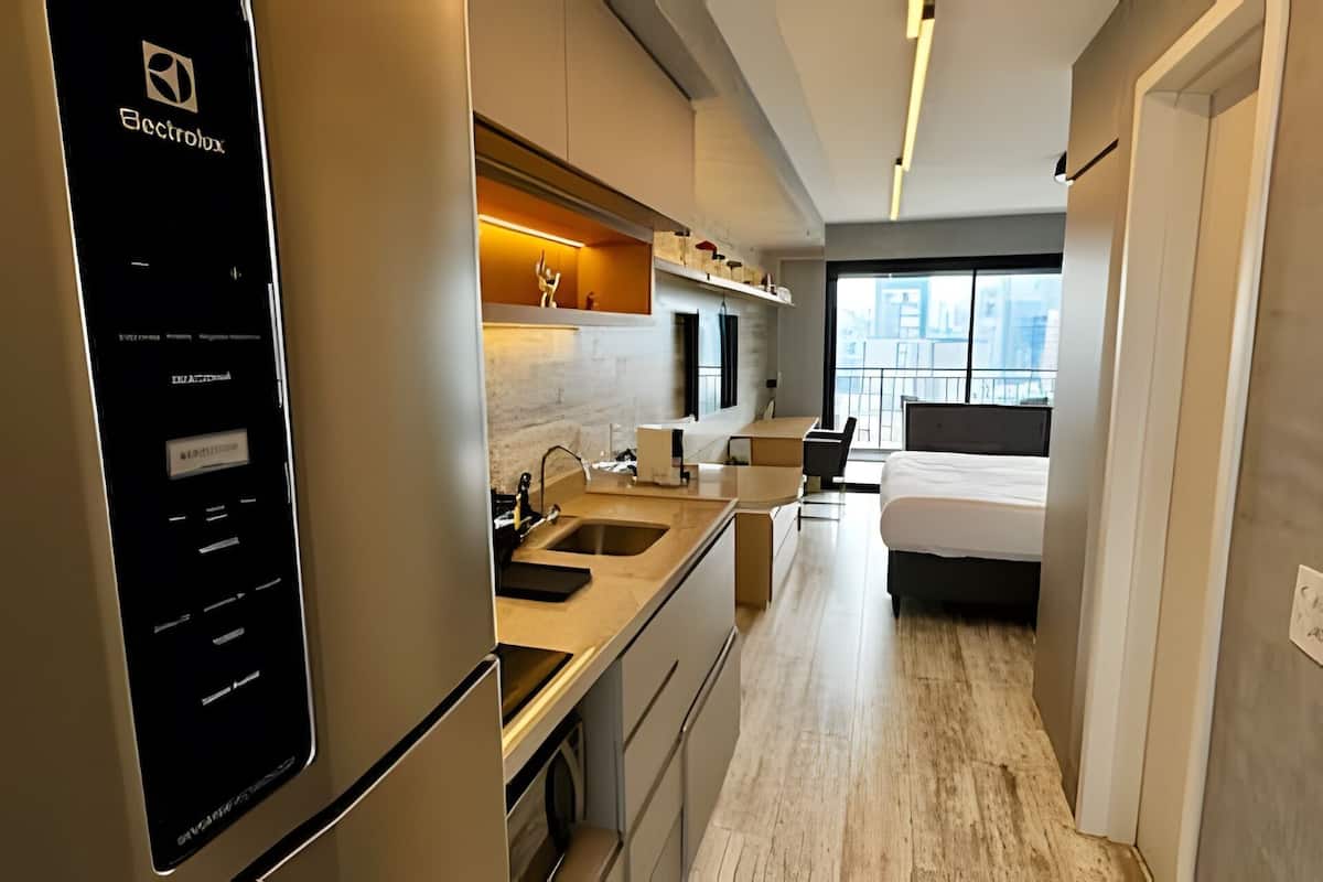 A modern hotel room with a kitchenette, a bed, a small seating area, and a view of the city.