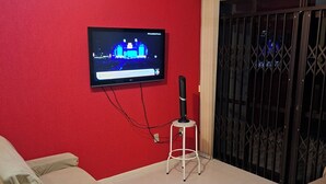 Un salon avec un mur rouge, un téléviseur à écran plat fixé au mur, un ventilateur de plafond, un canapé recouvert d’une housse, un petit tabouret blanc et une table sur laquelle se trouvent une paire de ciseaux et un tournevis.