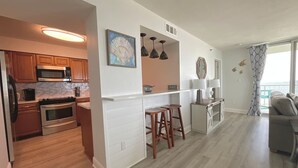 Una cocina moderna con armarios de madera, horno empotrado y microondas. Hay dos taburetes de madera y una pequeña isla con un fregadero. La cocina da a un comedor con vistas al océano.