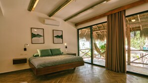Un dormitorio espacioso con una cama grande, suelo de madera y vistas a un espacio exterior con techo de paja y vegetación.