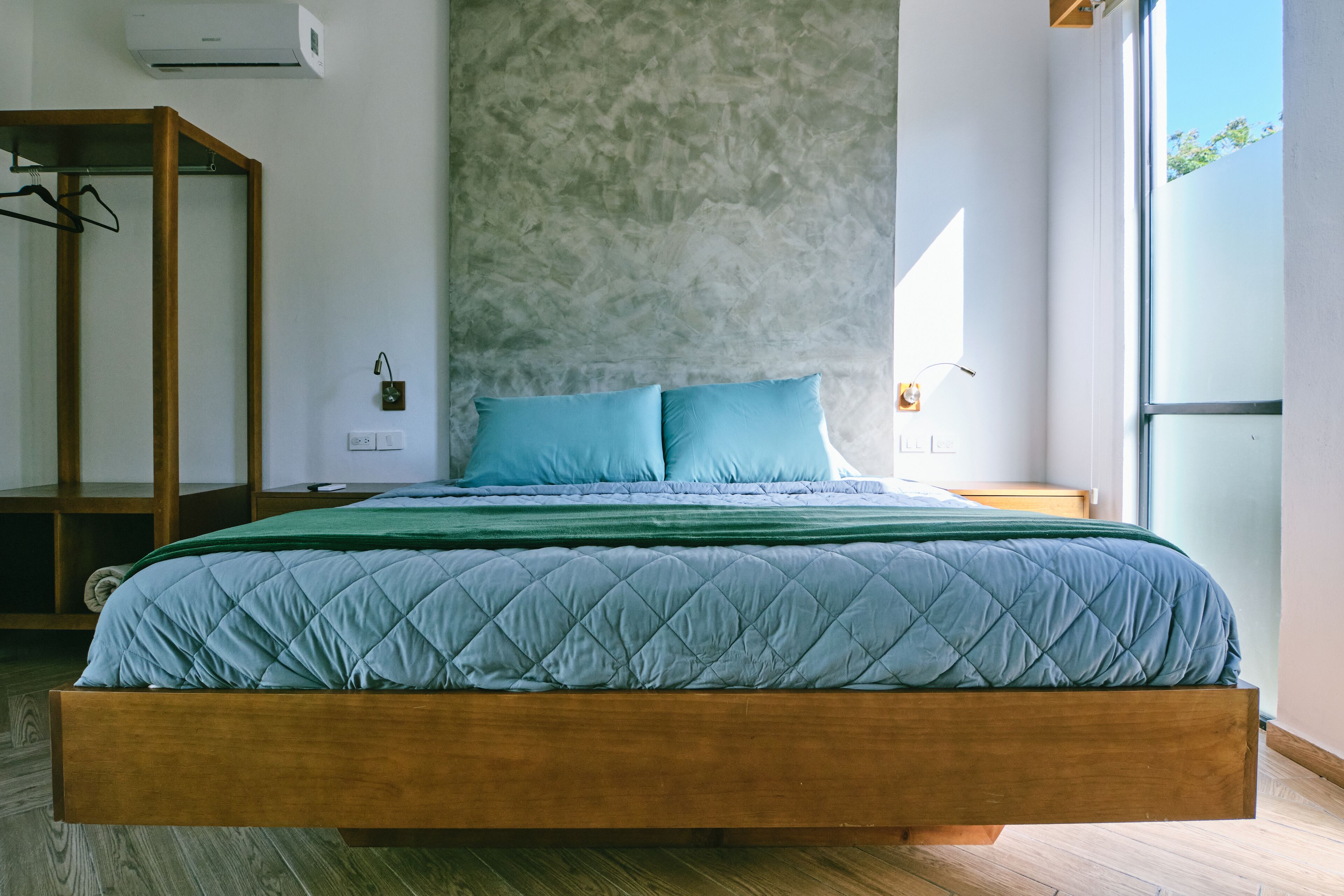 A modern bedroom with a wooden bed frame, a concrete accent wall, and a large window providing natural light.