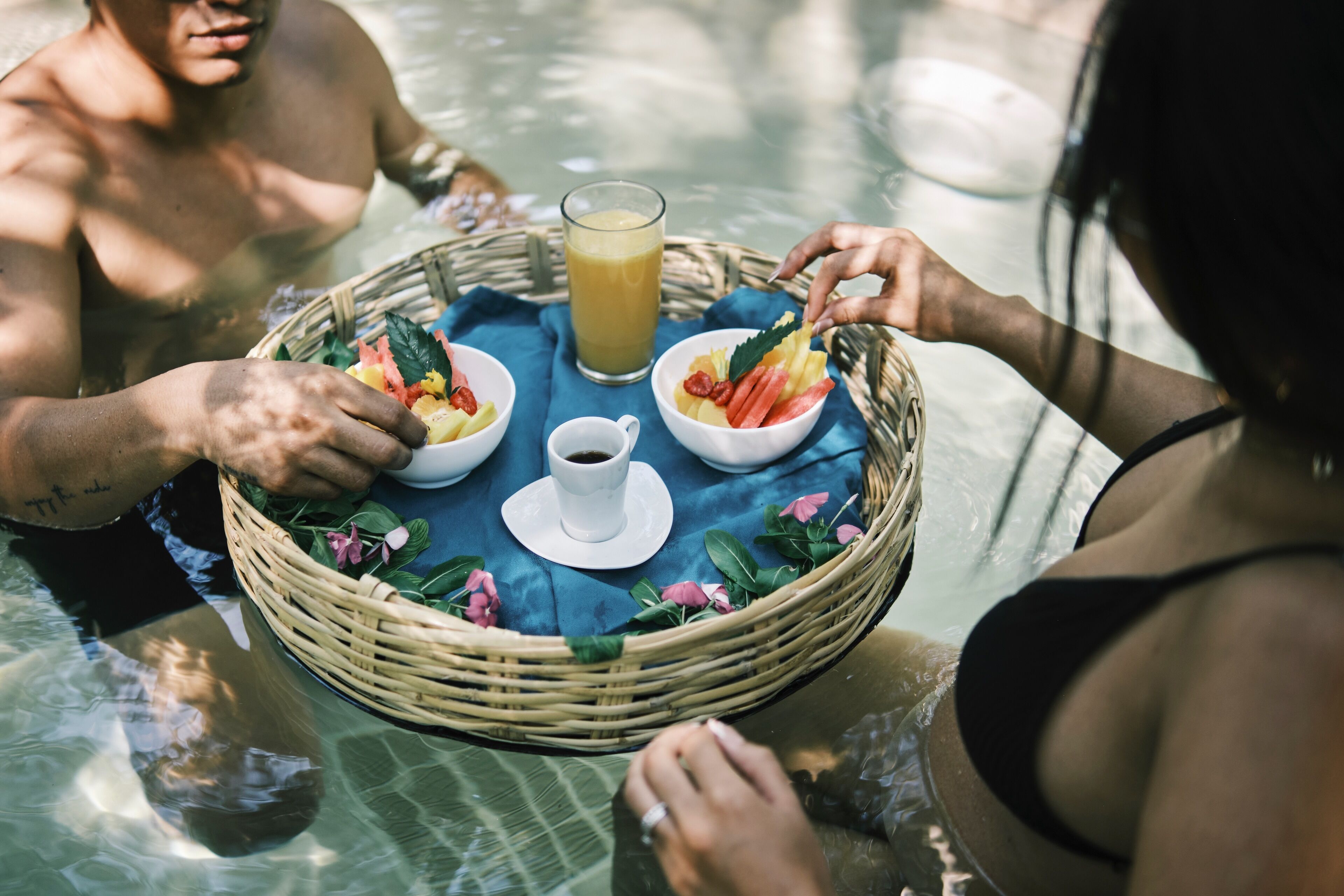 Una mesa de comedor con una cesta, tazones de comida y un vaso de jugo en un entorno junto a la piscina.
