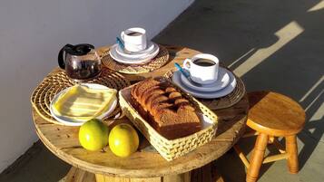 A round wooden table with a wicker placemat, two white cups with saucers, a metal coffee pot, a loaf of bread, and two lemons.