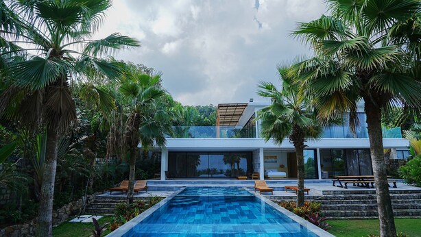 A modern pool area with a clear blue pool, surrounded by palm trees and a contemporary building with large windows. - Kiwuki Luxury Villa  (Phu Quoc)