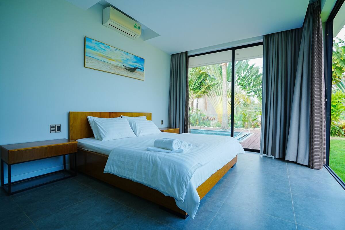 A modern hotel room with a large bed, a wooden nightstand, a painting, and a view of a pool and greenery.
