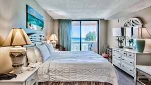 Habitación de hotel con una cama grande, dos mesitas de noche, un aparador y vista al océano.