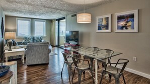 A modern living room with a sofa, a glass dining table with chairs, a TV, and artwork on the wall. - Beach Front, First Floor at Jetty East! One FREE Beach Chair Set Up Daily! (Destin)