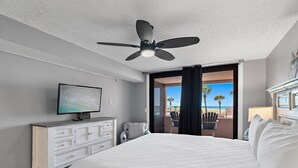 Habitació d'hotel amb un llit, una televisió, un ventilador de sostre, una petita estant i vista a la platja.