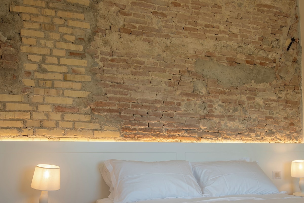 A bedroom with a brick wall, a wooden nightstand with a lamp, a bed with white bedding, and another lamp on the opposite side.