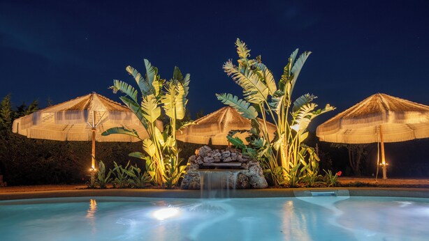 A pool with a waterfall feature, surrounded by thatched umbrellas and tropical plants. - Molikè Country House  (Giulianova)