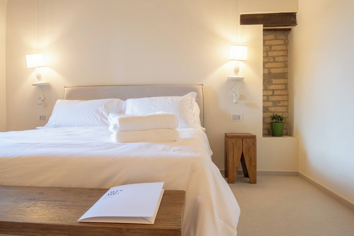 A neatly made bed with white linens, a wooden nightstand with a book, and a small green plant on a brick wall.
