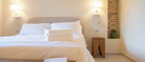 A neatly made bed with white linens, a wooden nightstand with a book, and a small green plant on a brick wall.