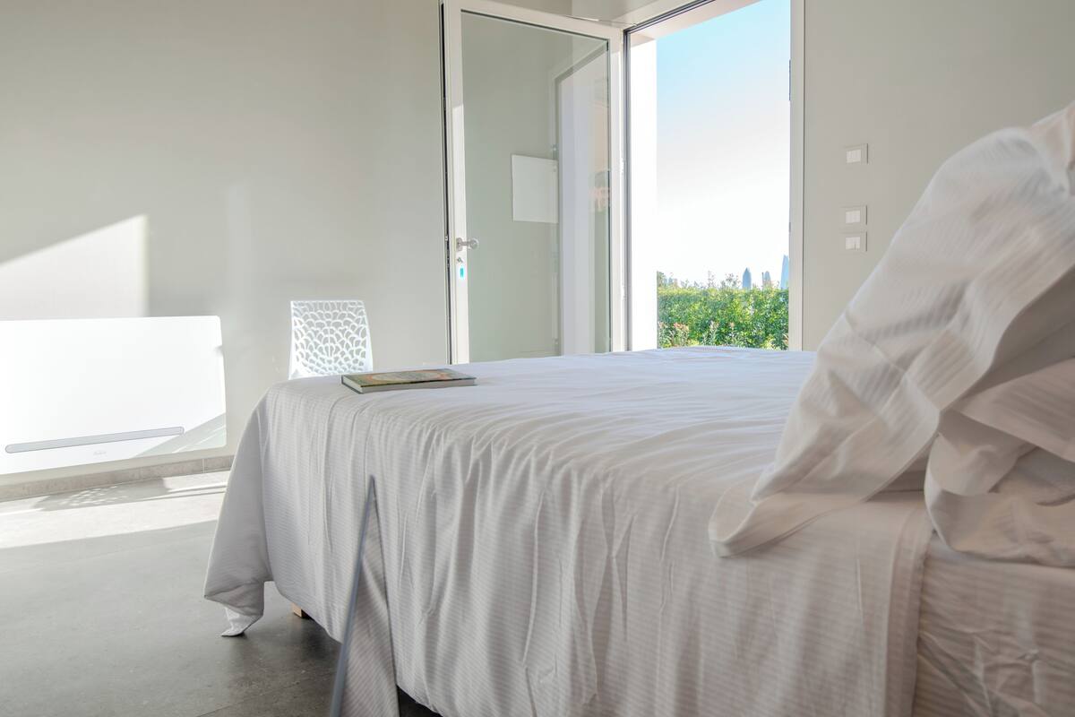 A bedroom with a large bed, a window with a view of greenery, and a door leading outside.