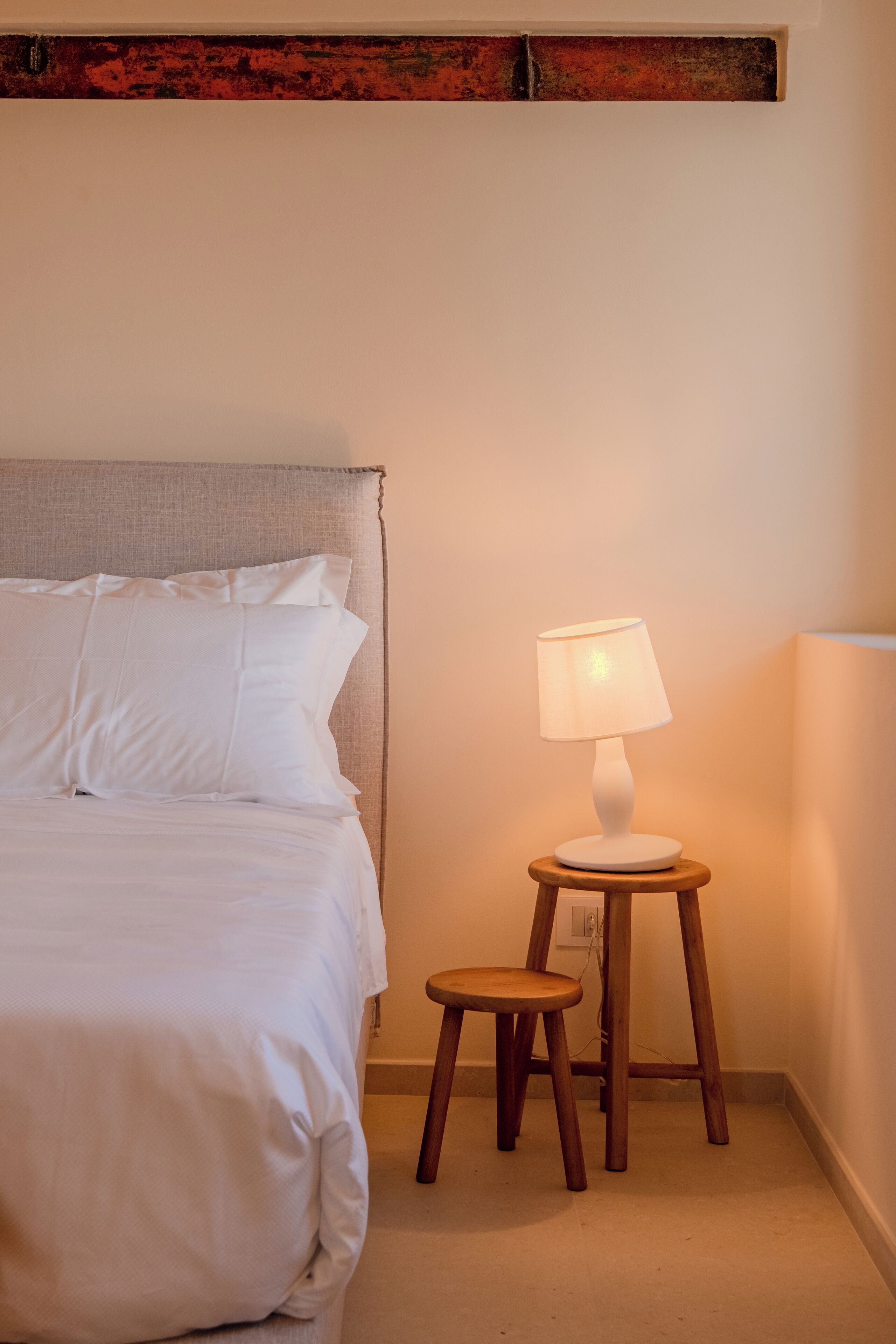 Una cama bien hecha con sábanas blancas, una mesita de noche de madera con una lámpara y un pequeño taburete de madera.