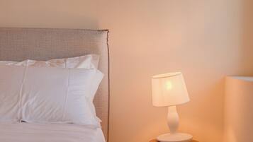 Una cama bien hecha con sábanas blancas, una mesita de noche de madera con una lámpara y un pequeño taburete de madera.