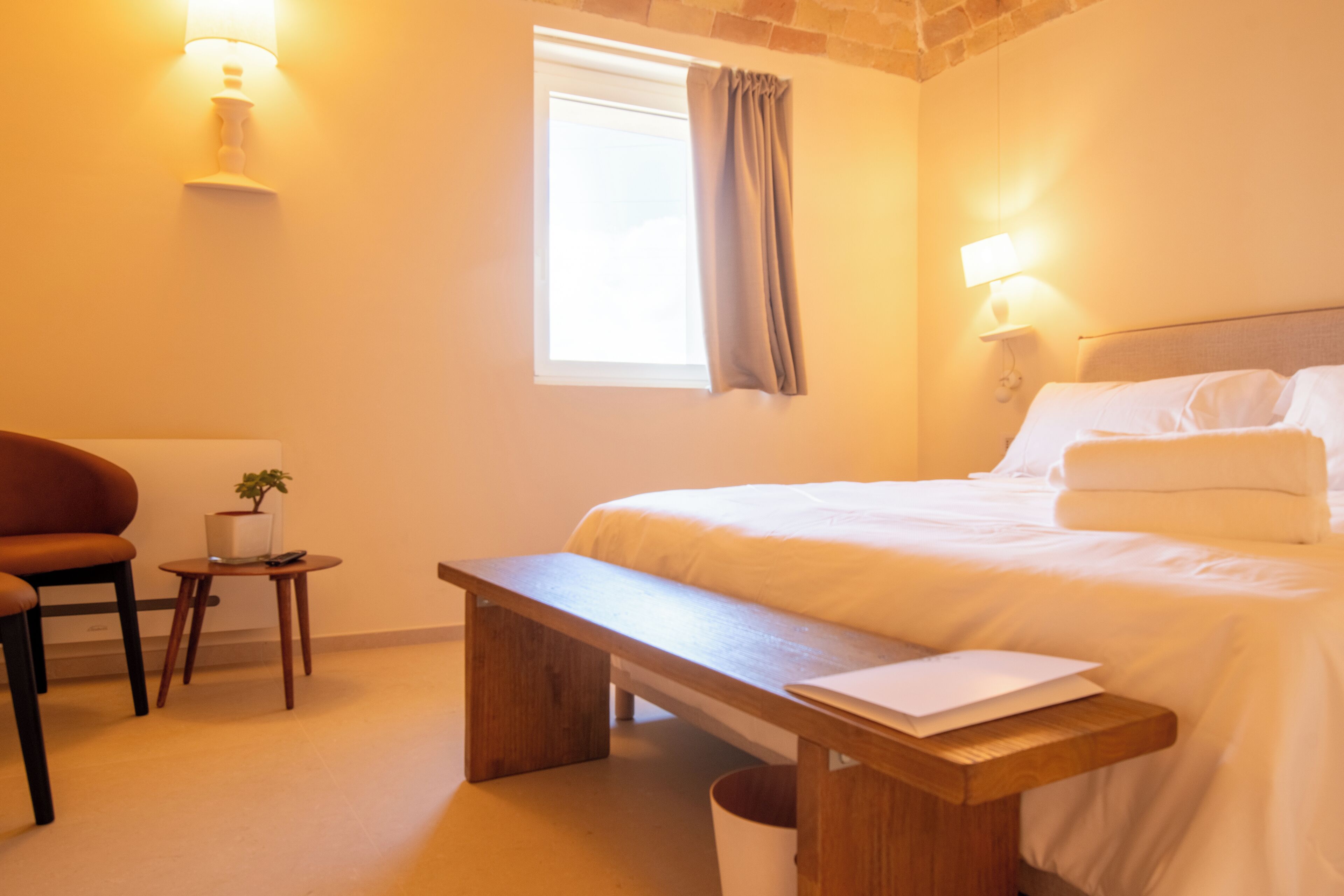 A hotel room with a bed, bedside table, lamp, window with curtains, and a small table with a plant.