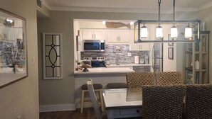 A modern kitchen with white cabinets, a built-in microwave, and a stainless steel refrigerator. There is a dining area with a table and chairs, and a decorative wall piece above the sink. - Beautifully Decorated Beach Front at Jetty East! One FREE Beach Chair Setup Dail (Destin)