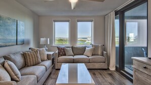 A modern living room with a beige sofa, a coffee table, and a ceiling fan. There are two windows with a view of the outdoors. - Newly Renovated December 2022 Beach Front at Jetty East! One FREE Beach Chair Se (Destin)