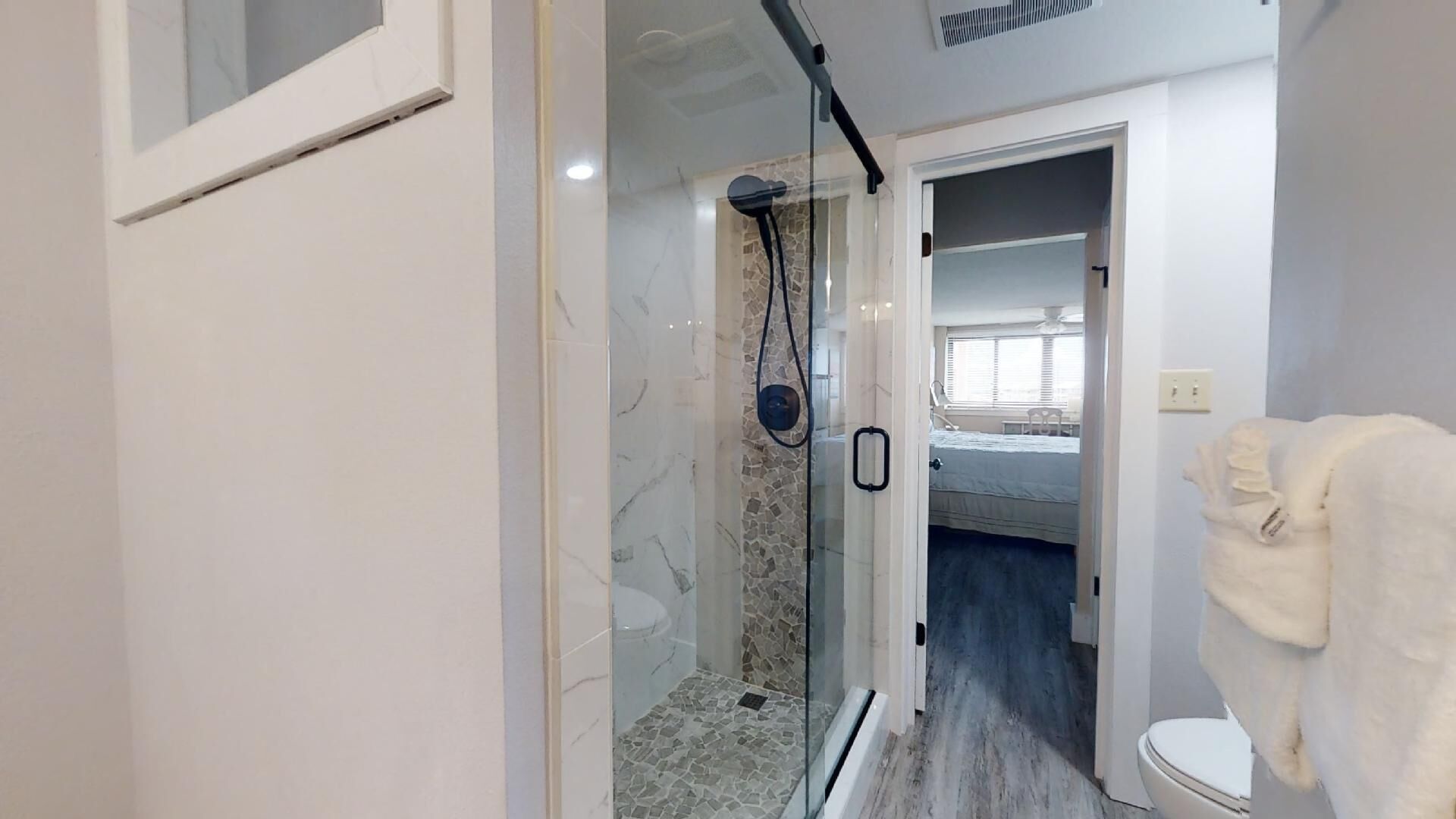 Ein modernes Badezimmer mit einer gläsernen Duschkabine, einer weißen Toilette und einem sichtbaren Schlafzimmer mit Bett und Fenster.