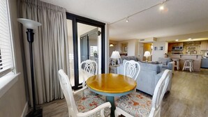 Un salon avec une table ronde en bois, des chaises blanches et un tapis à motifs. On aperçoit un canapé et un coin cuisine à l’arrière-plan.
