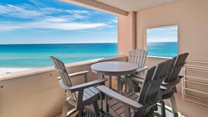 Un balcón con una mesa redonda y sillas, con vista a una playa de aguas azules y un cielo parcialmente nublado.