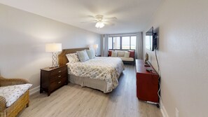 Un dormitorio con cama, mesitas de noche, una silla, un escritorio, ventilador de techo y televisor.