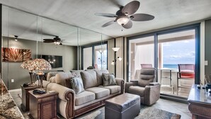 Ein geräumiges Wohnzimmer mit einer großen Couch, einem Couchtisch und einem Fernseher. Es gibt einen Deckenventilator, eine Lampe und einen Blick auf den Ozean durch große Fenster.