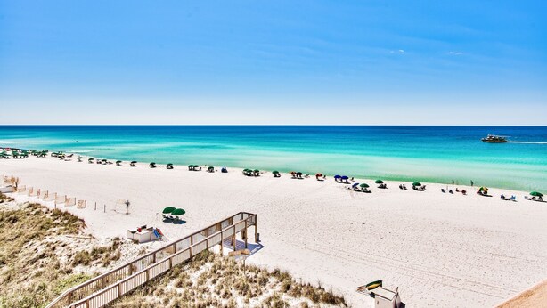 A beach with a wooden walkway leading to a structure, a clear blue sky, and a boat on the water. - Gulf view unit at Jetty East! One beach chair set up daily. (Destin)