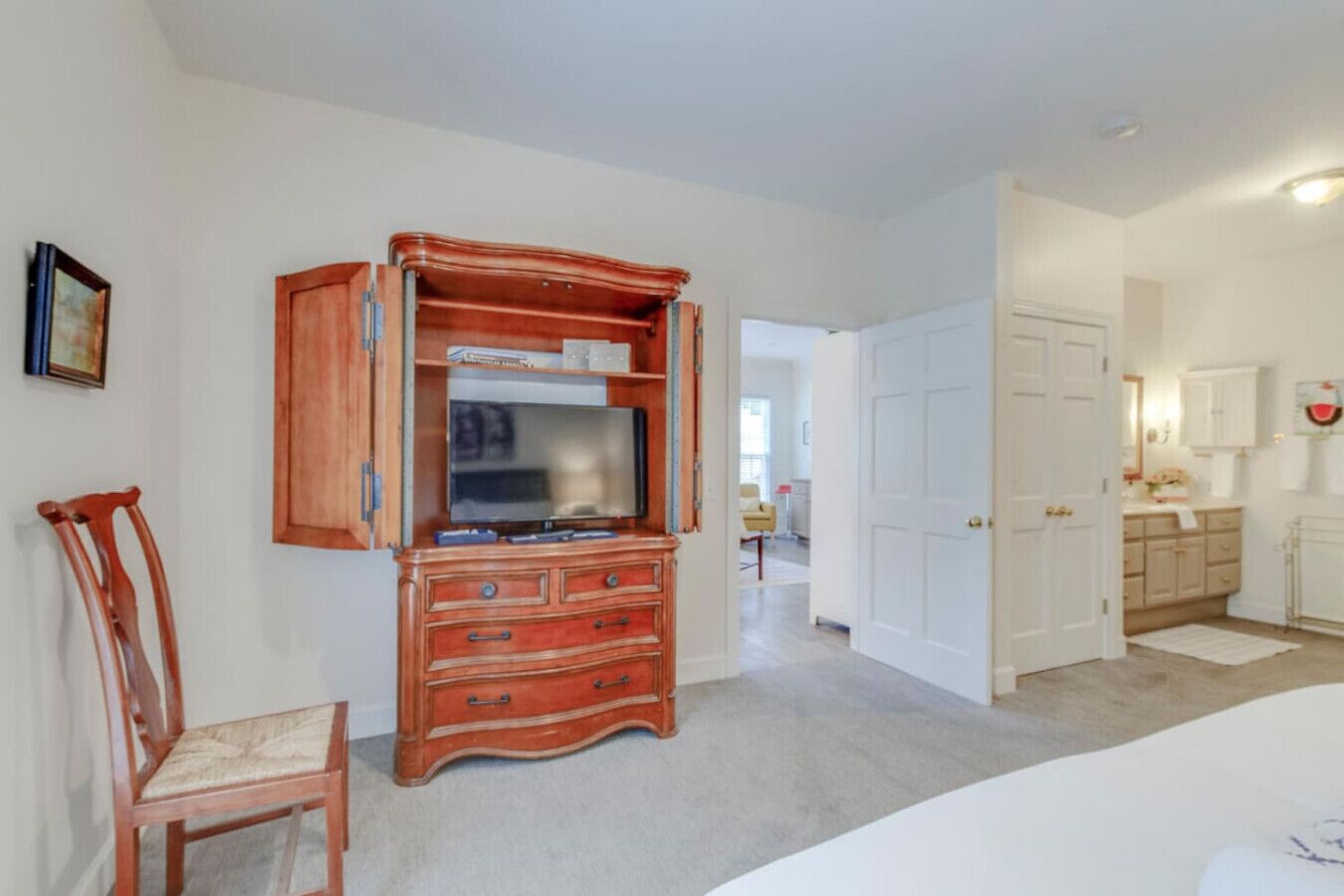 Une chambre avec une commode en bois, une télévision et une chaise. Un couloir mène à une salle de bain équipée de toilettes et d’un lavabo.