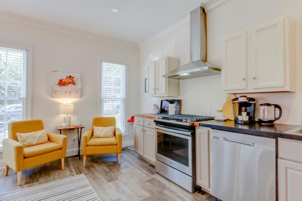 A modern kitchen with white cabinets, a stainless steel stove, and a coffee maker. There are two yellow chairs and a small table with a lamp.