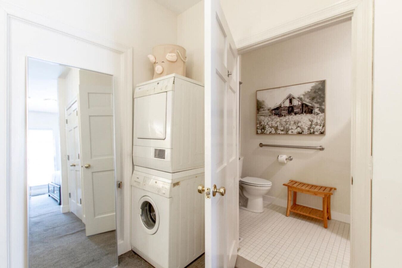 A laundry room with stacked washers and dryers, a toilet, a small wooden bench, and a wall-mounted towel rack.