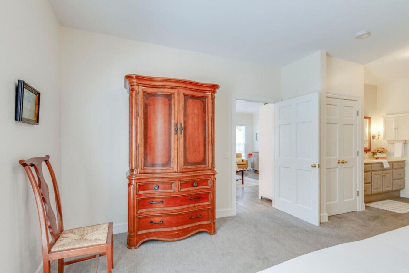 A bedroom with a large wooden armoire, a wooden chair, and a white door leading to a bathroom.