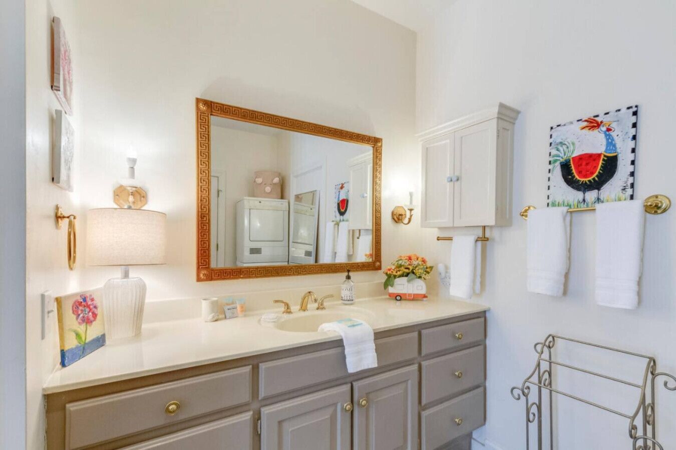 Un baño con un espejo grande, un lavabo, un mueble de tocador con cajones, un perchero para toallas y un cuadro decorativo en la pared.