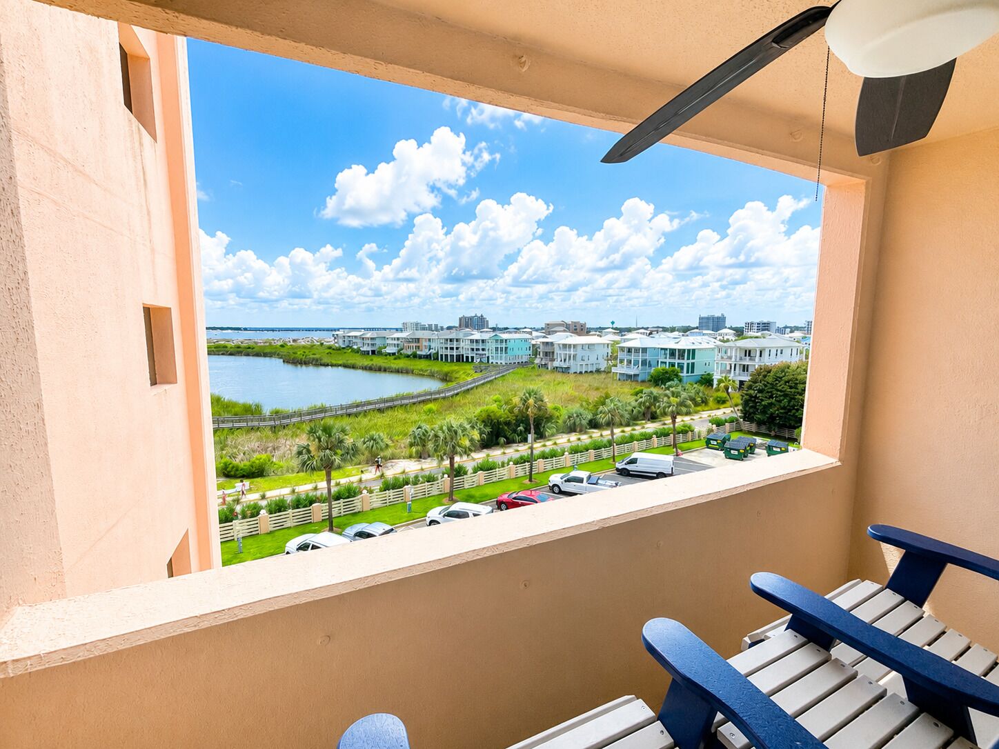 A balcony with a view of a waterfront, a paved walkway, and a row of parked cars.