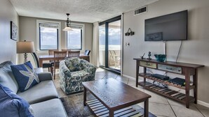 Ein Wohnzimmer mit einer Couch, einem Couchtisch und einem Fernseher an der Wand. Es gibt einen Essbereich mit Stühlen und einem Tisch.