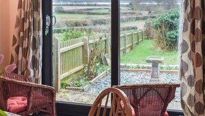 A room with a large window offering a view of a garden with a wooden fence, a stone bench, and a wicker chair. - 2 bedroom accommodation in Shotley, Ipswich (Shotley, Ipswich)