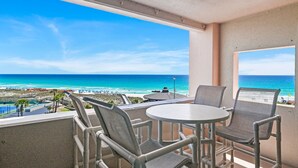 A balcony with two chairs and a table, overlooking a beach with clear blue water and a distant shoreline. - Spacious three-bedroom unit! One free beach chair set up daily. (Destin)