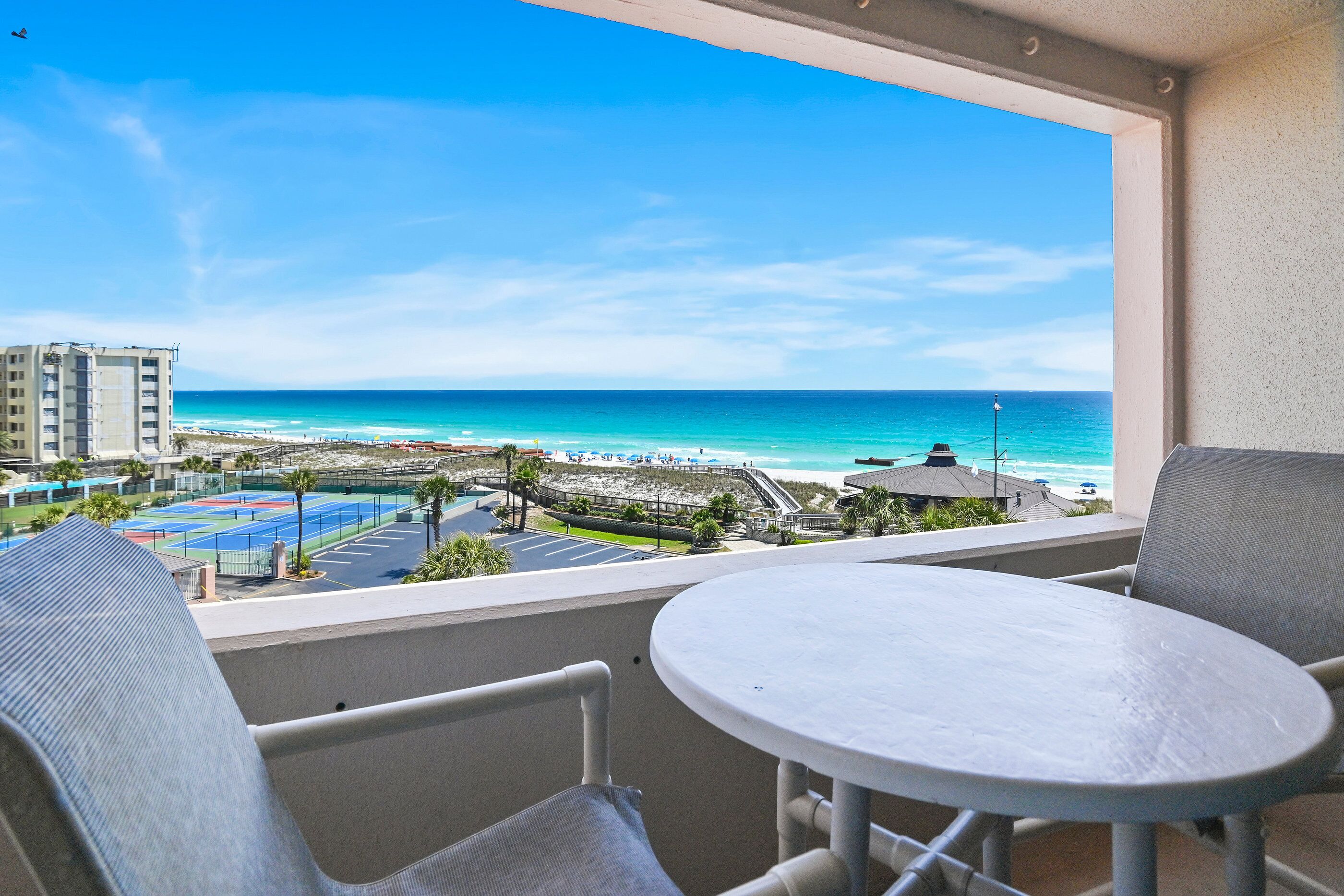 A balcony with a table and chairs, overlooking a beach with a tennis court and buildings in the distance.