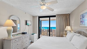 A hotel room with a bed, dresser, ceiling fan, and a balcony with a view of the ocean.