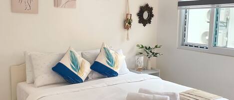 A neatly arranged bedroom with a bed, pillows, wall art, a mirror, and a window with blinds.