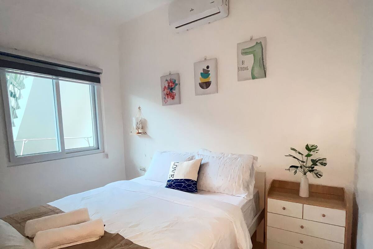 A bedroom with a bed, a bedside table, a nightstand, a window, and wall art.