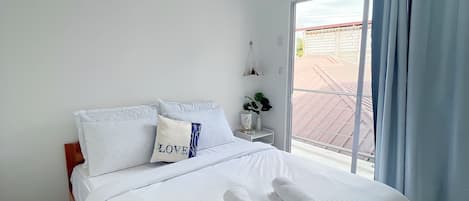 A neatly made bed with white linens and two rolled towels, a bedside table with a vase, and a view of a building through a glass door.