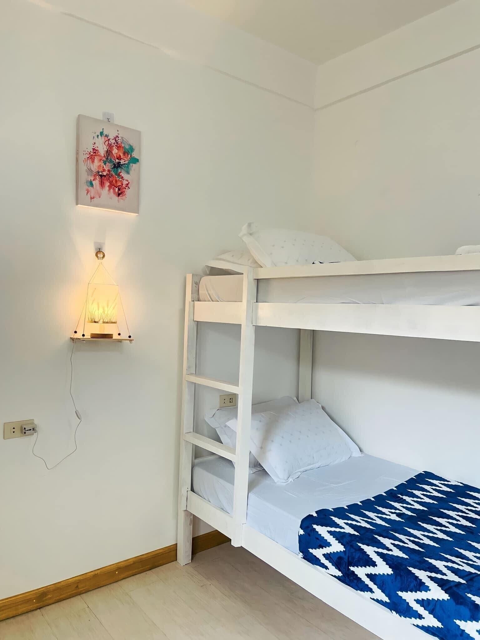 Un lit superposé avec une tête de lit blanche et une couverture bleue à motifs. Il y a un luminaire fixé au mur et un tableau encadré au-dessus du lit.
