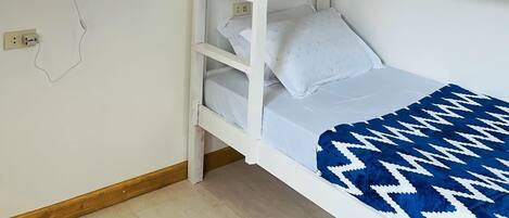 A bunk bed with a white headboard and a blue patterned blanket. There is a wall-mounted light fixture and a framed picture above the bed.