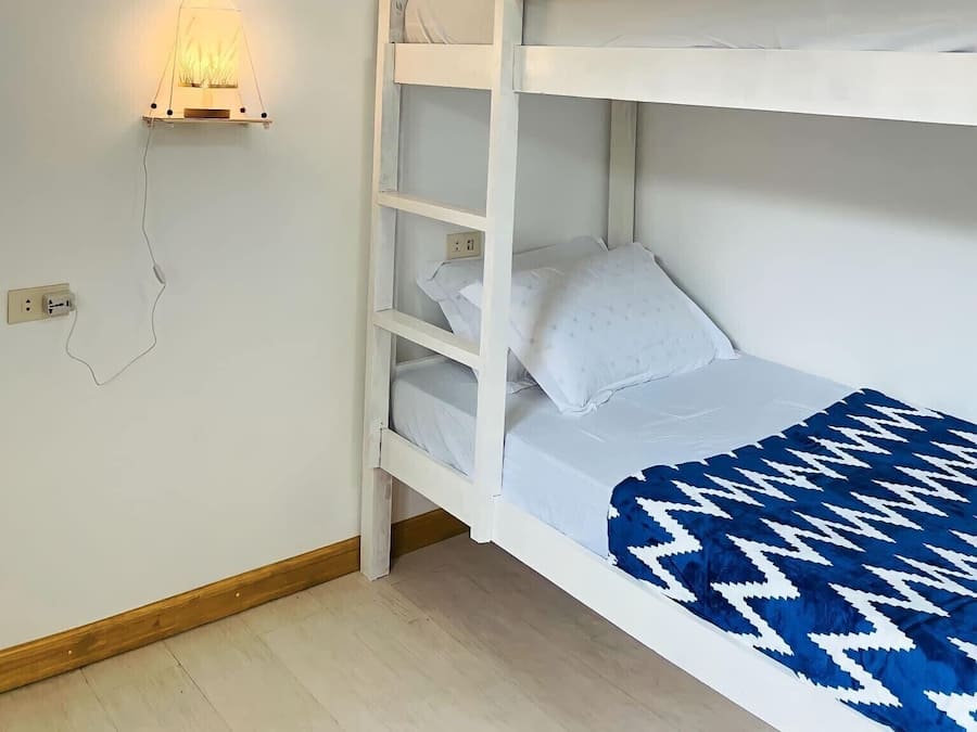 Una cama individual con cabecera blanca y manta estampada azul. Se observa un aplique de pared y un cuadro enmarcado sobre la cama.