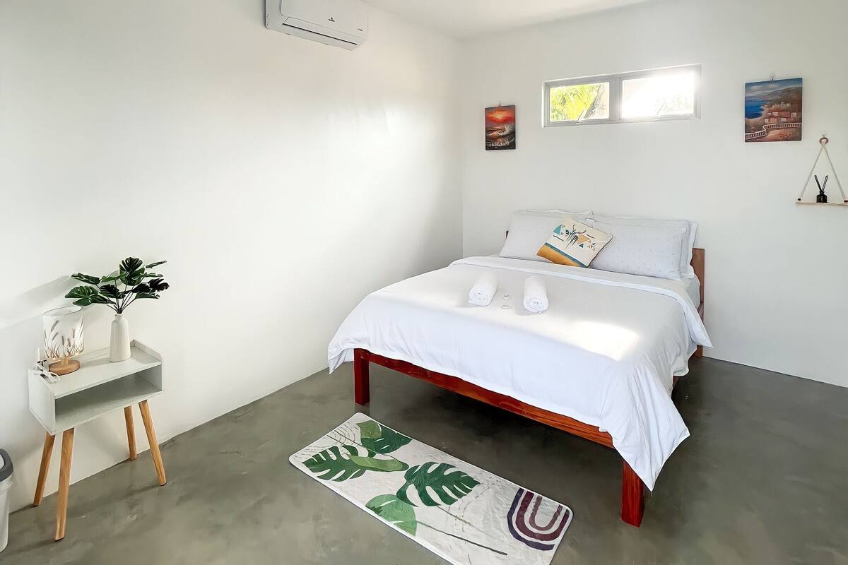 A minimalist bedroom with a bed, a small side table with a plant, and a concrete floor.