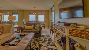 A living room with a sofa, armchair, and a coffee table. There is a flat-screen TV mounted on the wall, a fireplace, and a window with curtains.