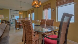 A dining area with wicker chairs and a glass table, large windows with blinds, and a hanging light fixture.