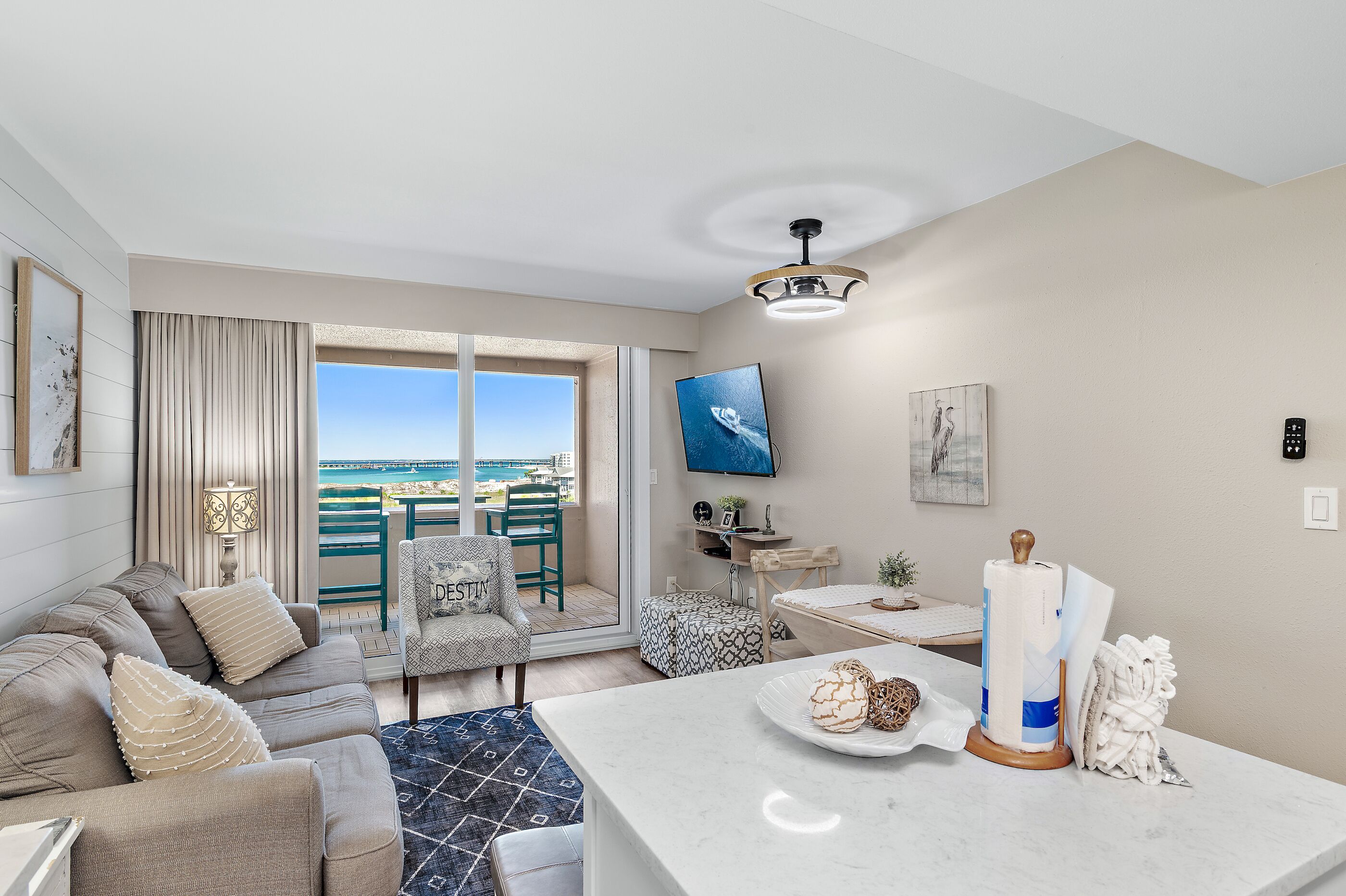A modern living room with a sofa, a dining area, a TV mounted on the wall, and a view of the sea through large windows.