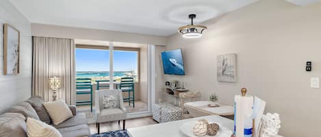 A modern living room with a sofa, a dining area, a TV mounted on the wall, and a view of the sea through large windows.
